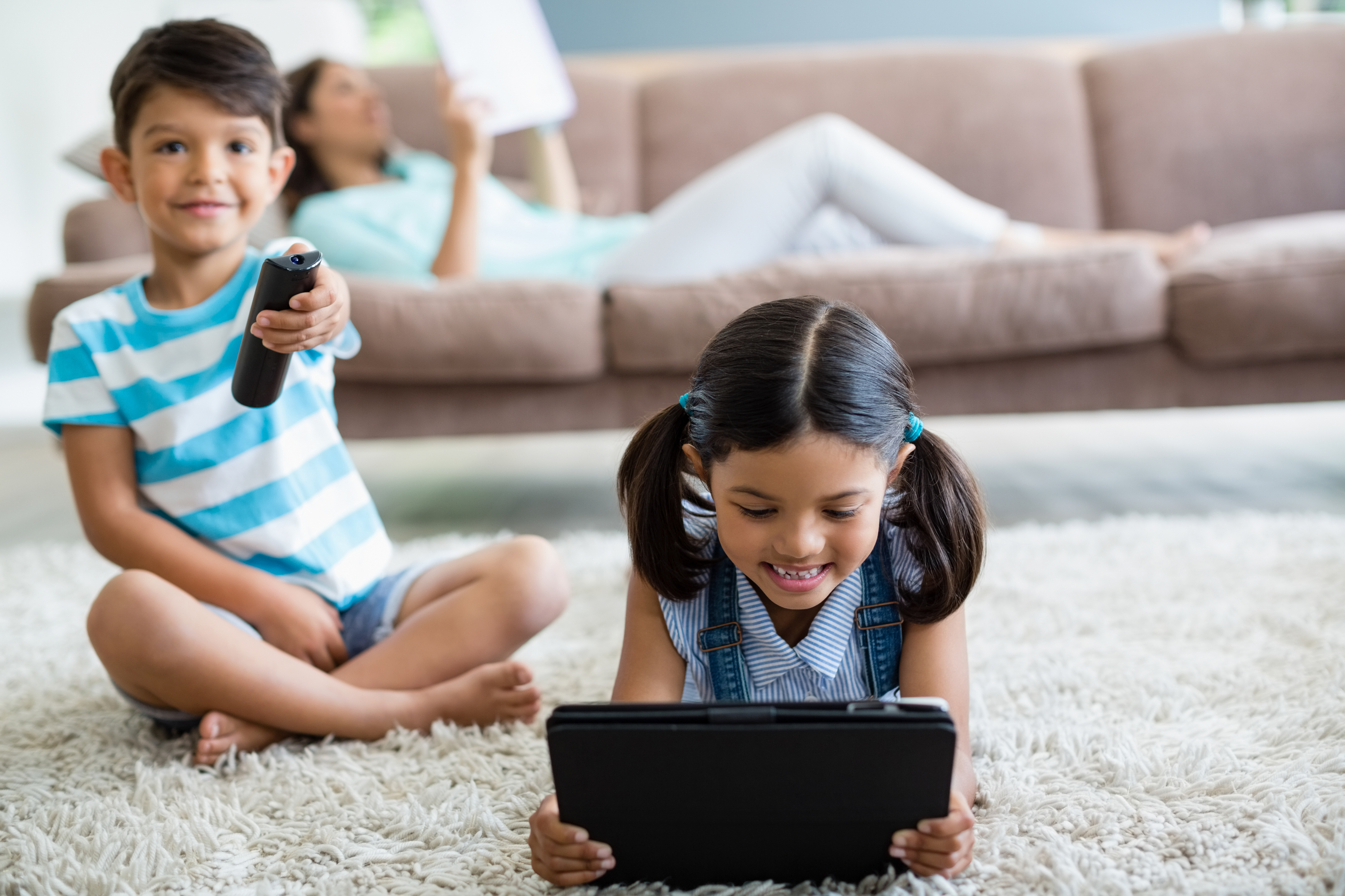 Image of a child holding a tv remote and another child holding an iPad - they are both sitting on the floor of their living room